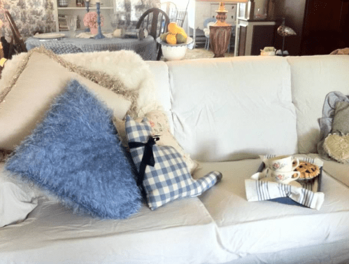 A warm and inviting photo of a cozy living room, featuring a beautifully made couch with a slip-cover, with plush pillows and a soft throw. In the background is a Bistro Area with a blue tablecloth, cottage styled Windsor chairs and a candle on the table. Two elegant lamps sit on either side of the couch, casting a soft glow over the room and creating a peaceful and relaxing atmosphere.