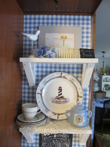 A cute coffee station scene with tiny white shelves against a blue and white gingham wallpapered wall and inviting amateur artwork of a beach scene above. The photo is of a warm and inviting spot in the kitchen of a cozy cottage, featuring a beautifully made coffee bar, and a coffee maker, with a vintage coffee cup and saucer beside the percolator coffee machine. There is an adorable tiny white picket fence encircling a seagull figurine and a plate with a lighthouse in the center. A candle is casting a soft glow over the vignette, creating a peaceful and relaxing atmosphere. Cottage style is featured. French Country home decor is the hope, relaxation is the activity. Comfortable living in a cozy home is the theme.